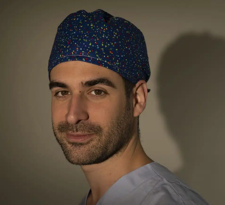 Man in blue speckled scrub cap and light blue top against beige wall, symbolic image for Mommy Makeover at BONITAS in Nuremberg