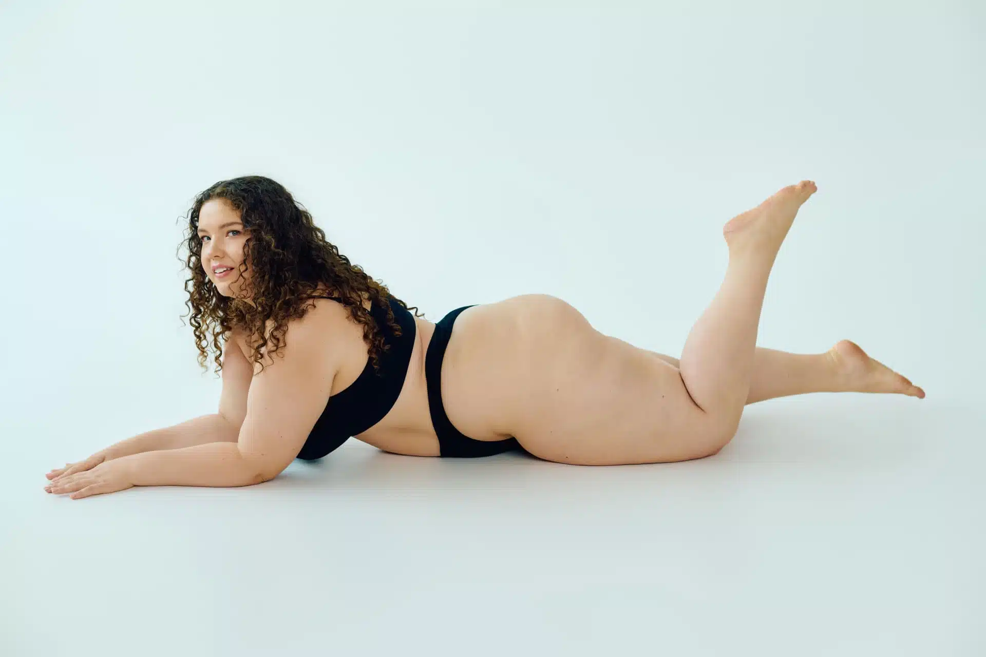 Woman with curly hair in black lingerie lying on her side on white background smiling, symbolic image for Lipedema Stage 3 at BONITAS in Nuremberg