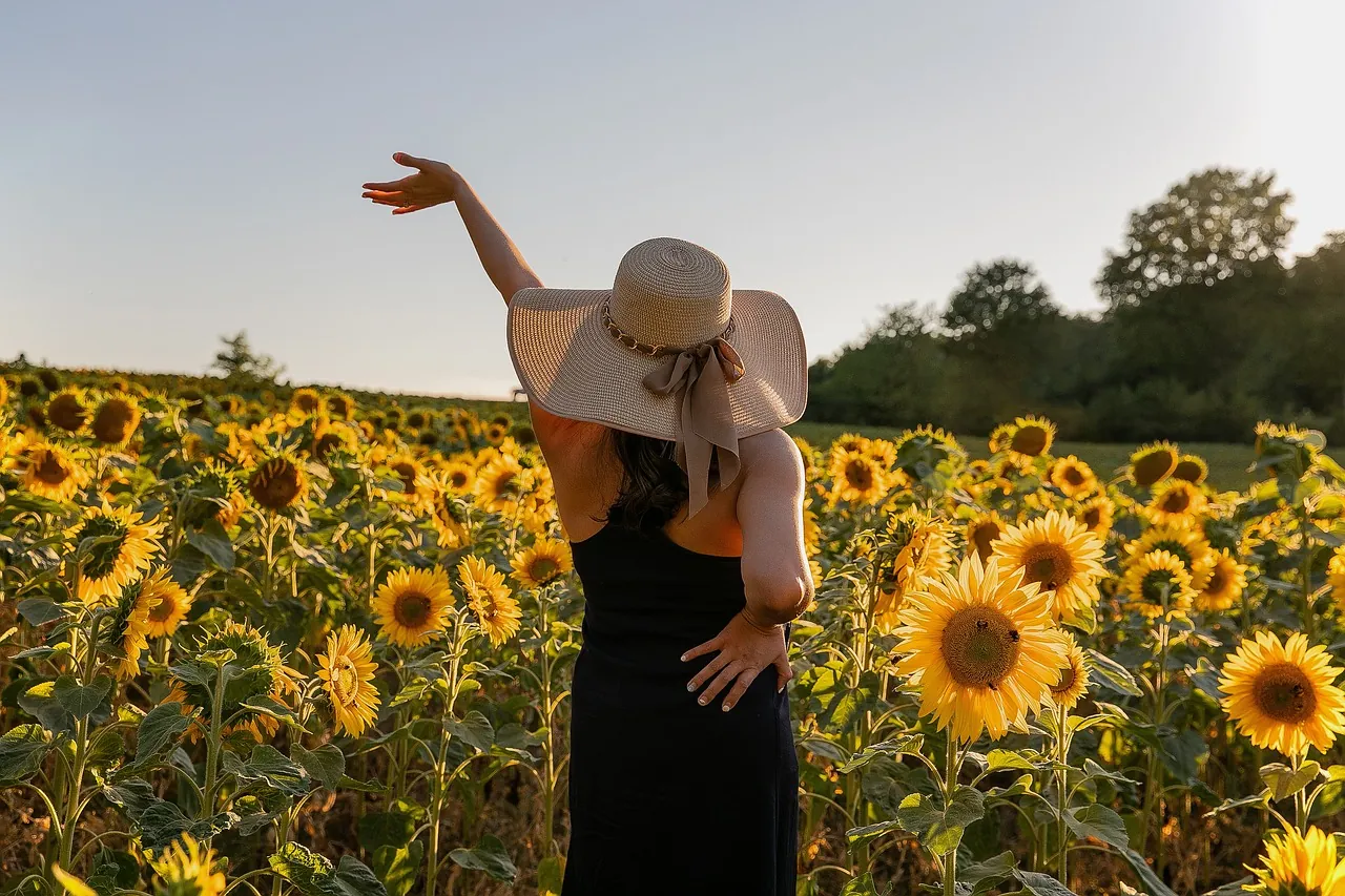 Mujer con sombrero de paja y vestido oscuro en un campo de girasoles con un brazo en alto, imagen simbólica sobre el síndrome de Poland en BONITAS Núremberg