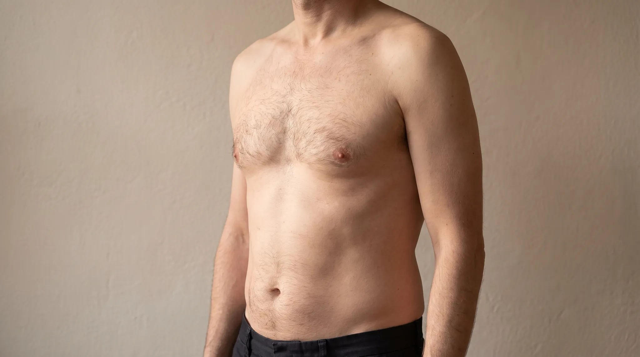 Upper body of a man in three quarter view with a flat chest against a beige wall, symbolic image on the subject of gynecomastia at BONITAS in Nuremberg