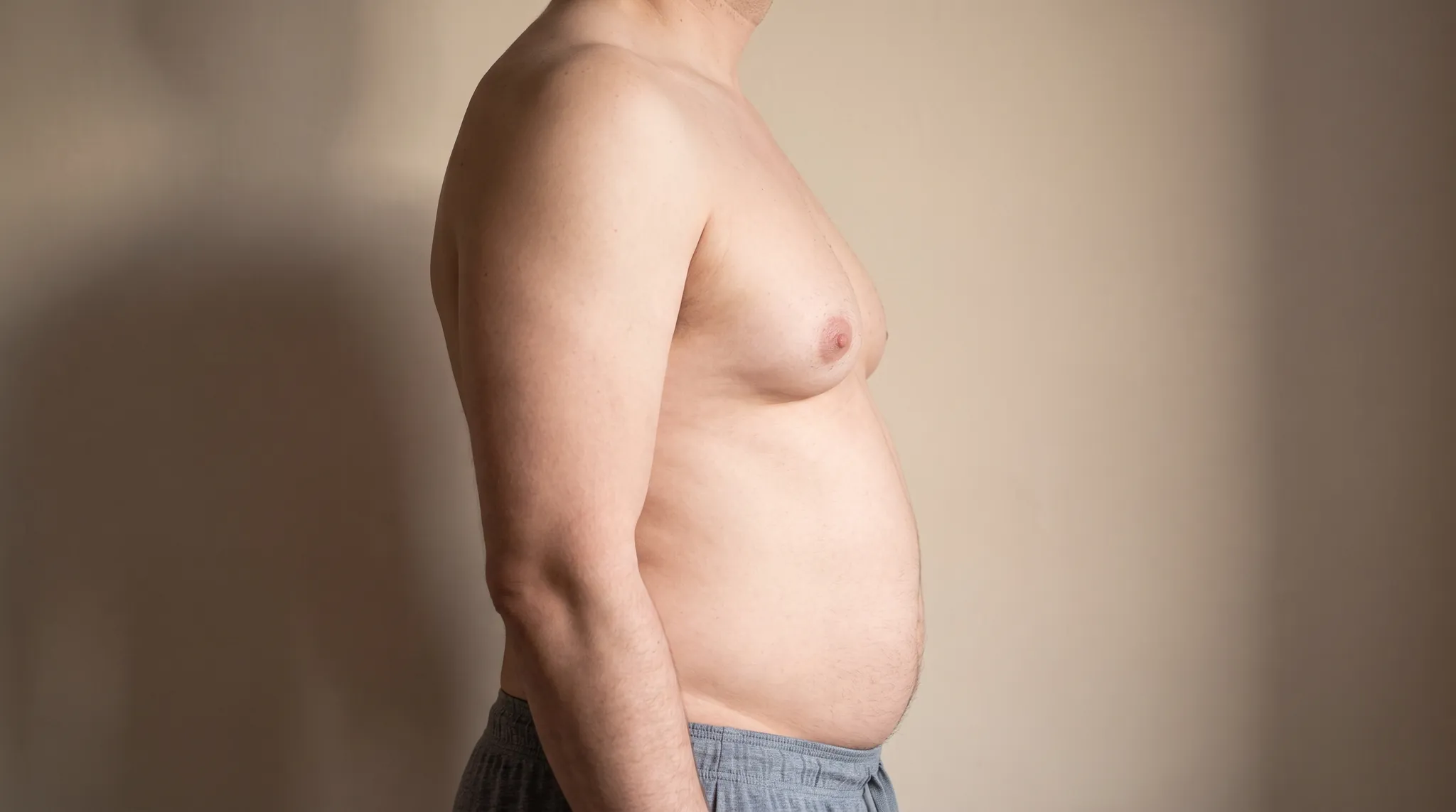 Side view of a mans upper body with the chest tissue projecting forward against a beige wall, symbolic image on the subject of gynecomastia without surgery at BONITAS in Nuremberg