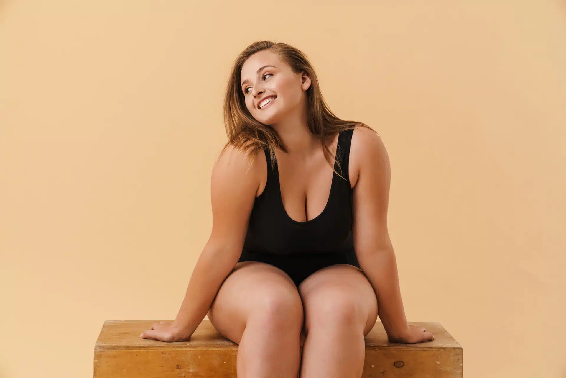 Mujer sonriente en body negro sentada en caja de madera ante pared beige, imagen simbólica sobre Liposucción para el lipedema en BONITAS Núremberg