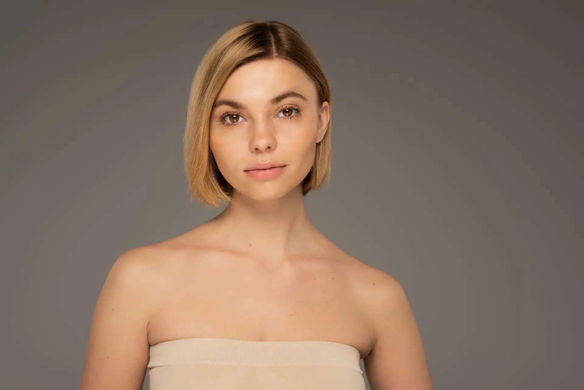 Mujer con cabello rubio bob en top beige sin tirantes frente a fondo gris, imagen simbólica sobre Elevar las comisuras de los labios en BONITAS Núremberg