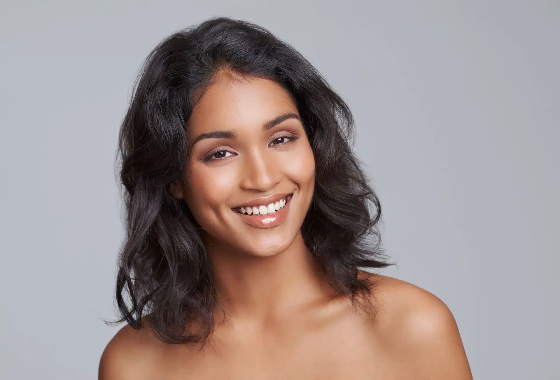 Mujer sonriente con cabello ondulado oscuro y hombros desnudos fondo gris, imagen simbólica sobre Relleno de ojeras en BONITAS Núremberg