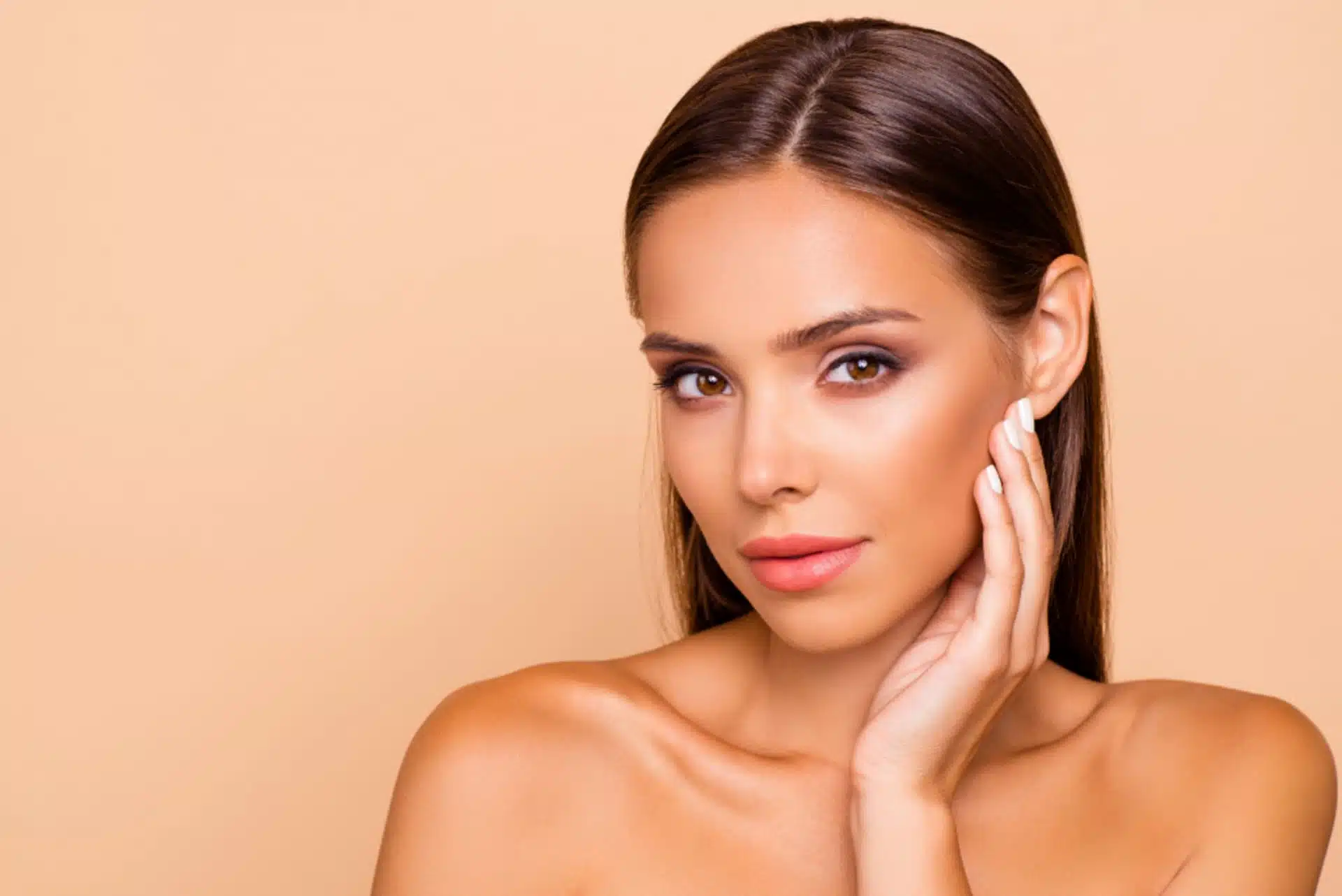 Woman with long brown hair touching her cheek against beige backdrop, symbolic image for Cheek Augmentation at BONITAS in Nuremberg