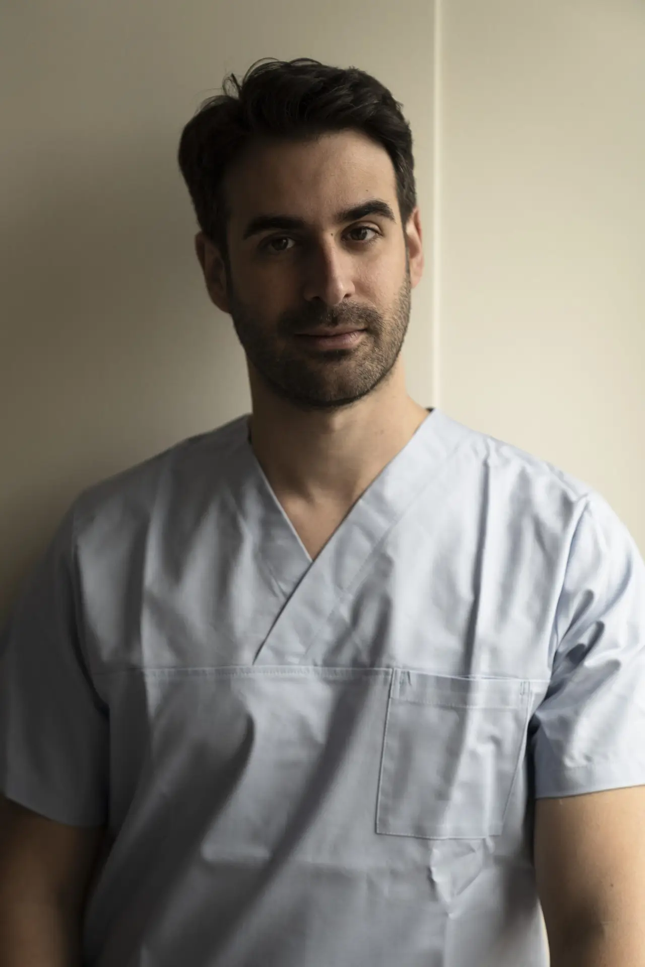 Mann in hellblauem Kasack lehnt an beiger Wand: Alejandro Marti, Facharzt für Plastische und Ästhetische Chirurgie, BONITAS Nürnberg