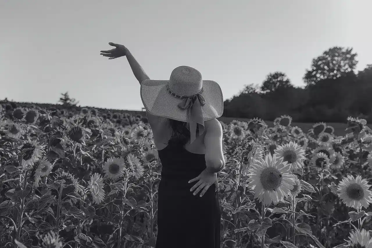 Mujer con sombrero de paja y vestido oscuro en un campo de girasoles con un brazo en alto, imagen simbólica sobre el síndrome de Poland en BONITAS Núremberg