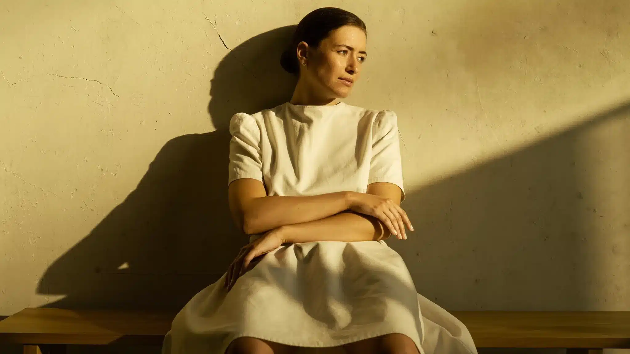 Mujer con vestido midi color crema, sentada en un banco de madera, los antebrazos apoyados ante la cintura, ante una pared beige, imagen simbólica sobre cuello alto y manos en el abdomen ante pared beige, imagen simbólica sobre Diástasis de rectos en BONITAS Núremberg